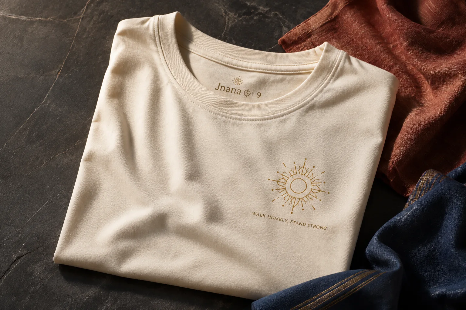 Cream Jnana 9 shirt concept with the message Walk humbly. Stand strong., alongside colored textiles